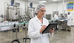 QA technician reviewing data on a tablet in a regulated manufacturing plant with filling and packaging equipment behind her.