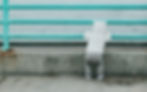 A baby stood up holding onto a rail looking out onto water. The baby's back is facing us and they are in a white hooded jacket with ears on (the hood is up), grey bottoms and baby trainers. The rail is meta, and turquoise in colour. The baby is stood on concrete. 