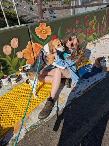 Artist Ana Gabriela sitting with dog in front of the Flower Bridge mural in Vancouver, Washington, featuring bright orange, yellow, and pink flowers along the bridge wall.”