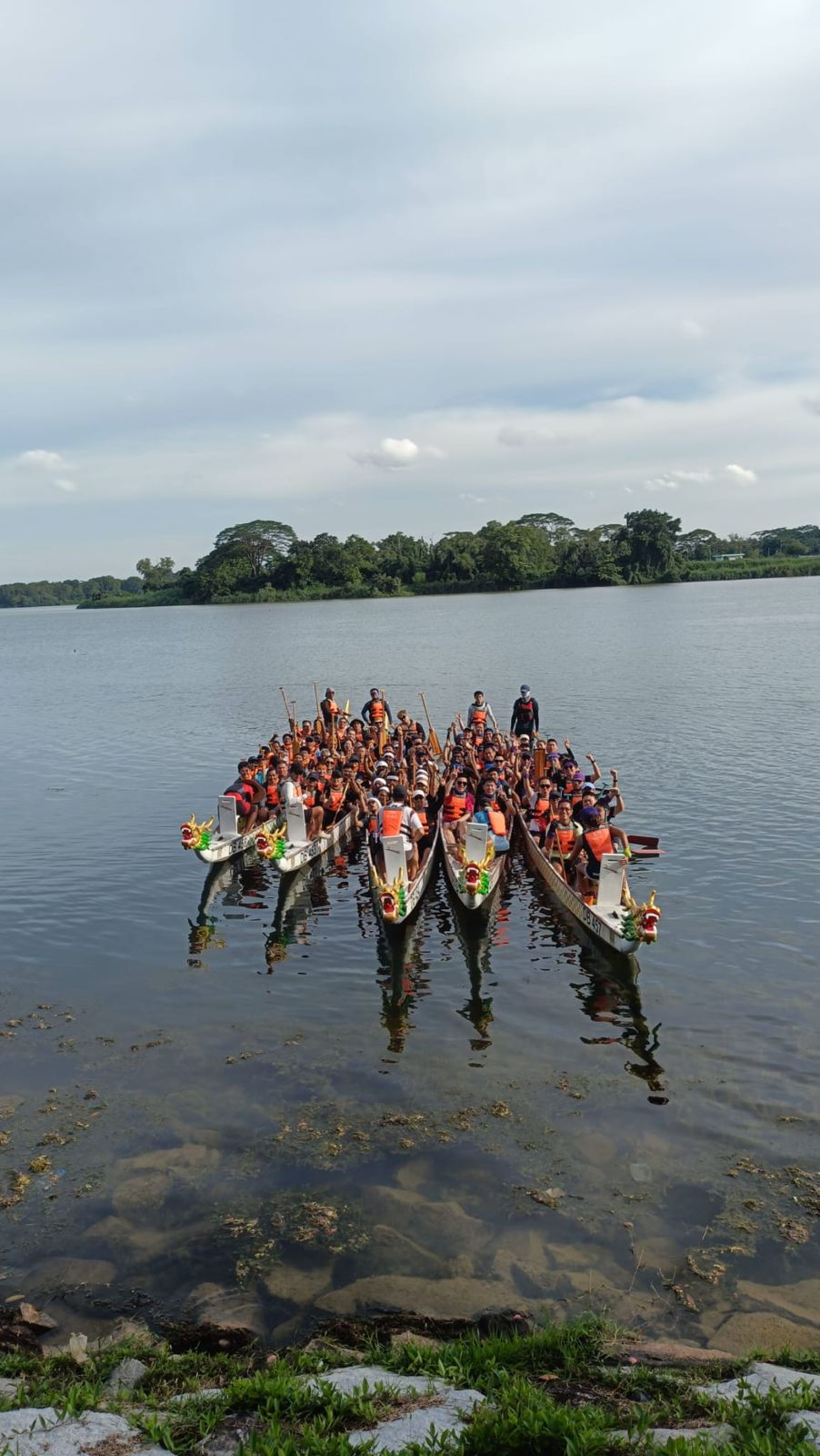 Emcee - The dragon boat racers of Safran prepared for a thrilling practice session on the serene lake with each team donning life jackets and paddles, ready to synchronize their efforts under the clear sky.