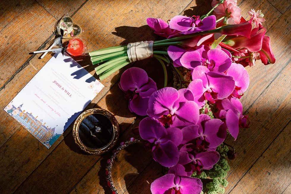 Pink orchids and calla lilies beside a JODIE & WILL wedding invite, ring box, and heart lollipops on a sunlit wooden floor.