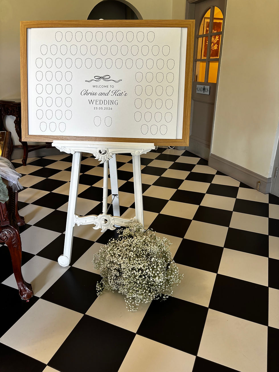 Wedding seating chart with blank face placeholders, on an easel beside baby’s breath on a black-and-cream checkered floor.