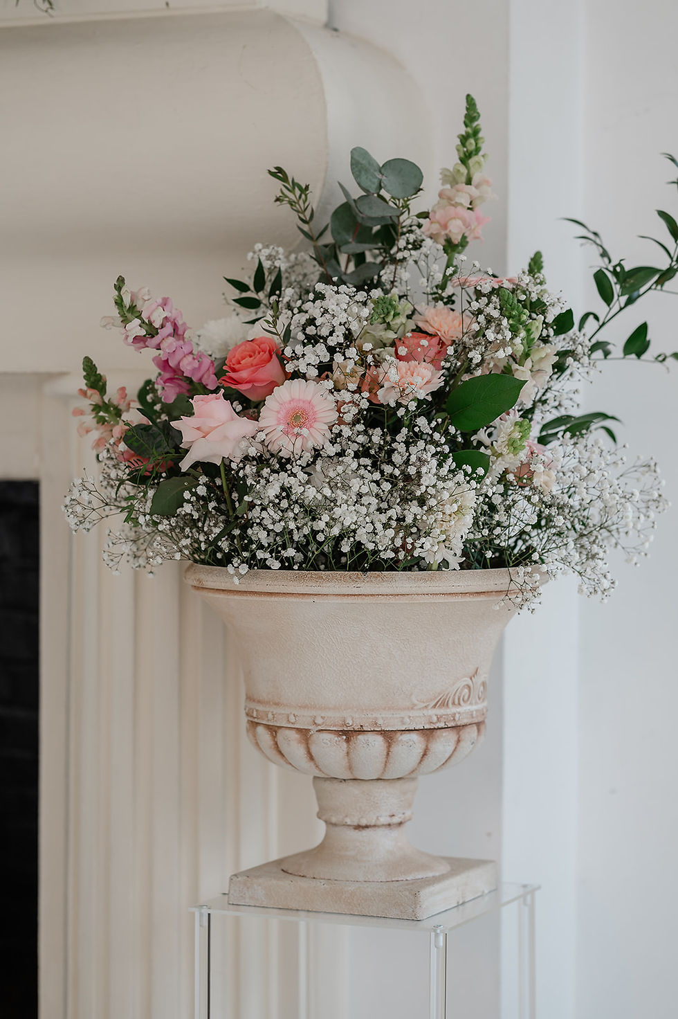 Pink and white floral arrangement in a beige urn on a stand beside a fireplace in a bright, elegant room at Northbrook Park.