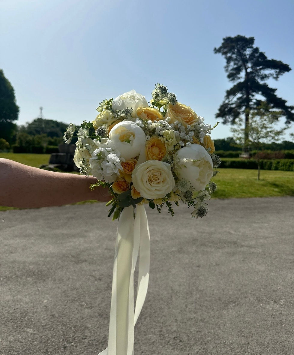Hand holding white and pale yellow rose bouquet with ribbon in a sunny park under a blue sky.