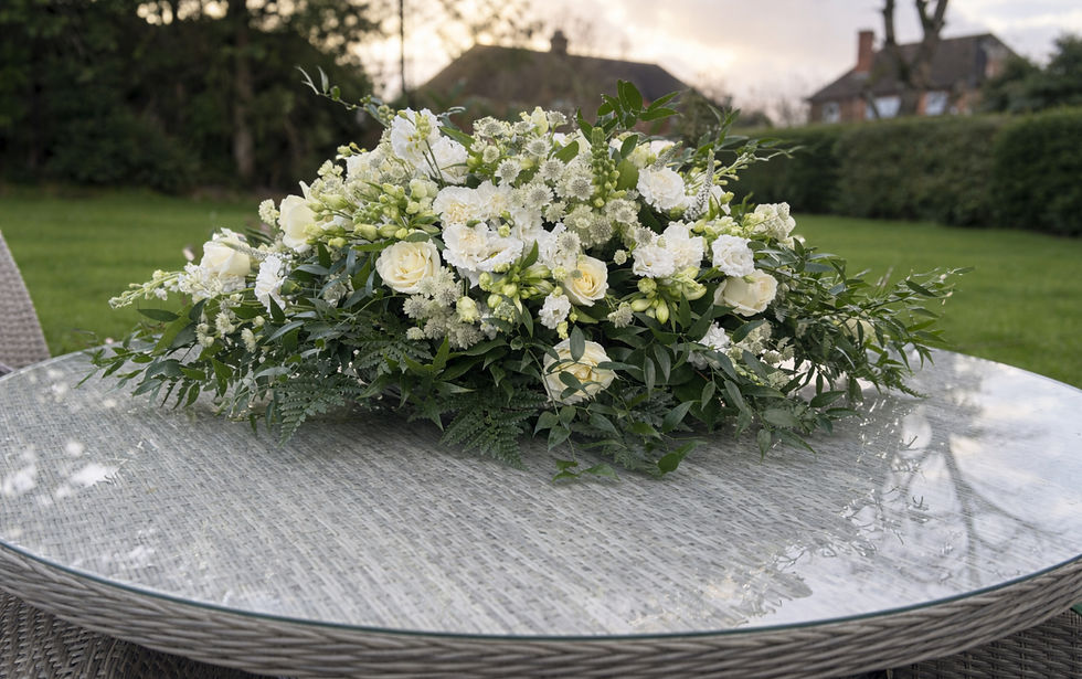 White rose and greenery casket spray on a glass patio table in a garden at dusk, with blurred houses in the background, in Alton, Hampshire