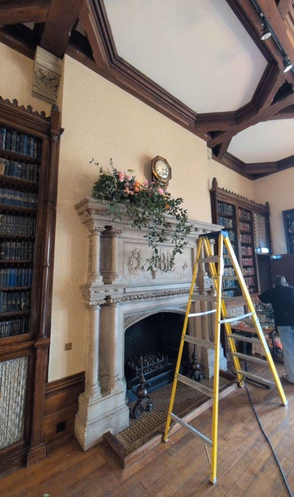 Yellow ladder in an ornate wood-paneled library at the elvetham hotel in hook beside a carved stone fireplace topped with flowers and a clock.