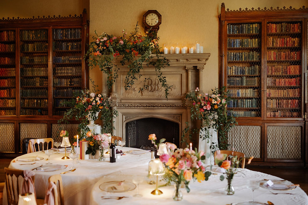 Elegant dining room with floral arrangements on a table by a fireplace. Bookshelves in the background and candles create a cozy ambiance.
