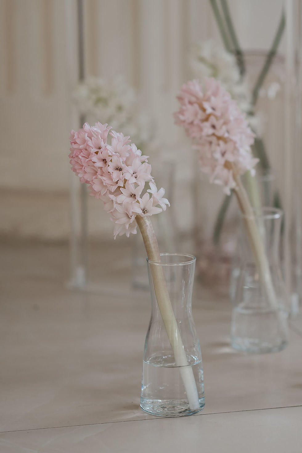 Pale pink flowers in clear glass vases on a beige table, with a softly blurred background and a calm, elegant mood.