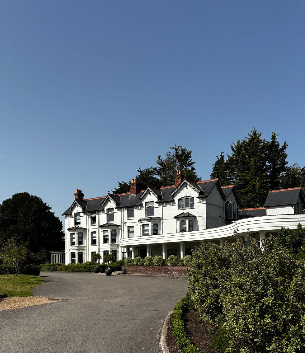Southdowns Manor a White mansion with black roofs and chimneys beside a driveway, framed by trees and shrubs under a clear blue sky.