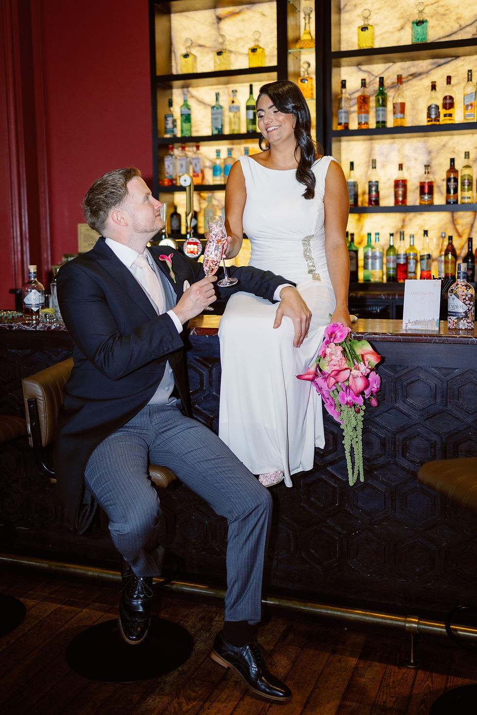 Bride in white dress and groom in suit toast with champagne at a bar, smiling before glowing liquor shelves.