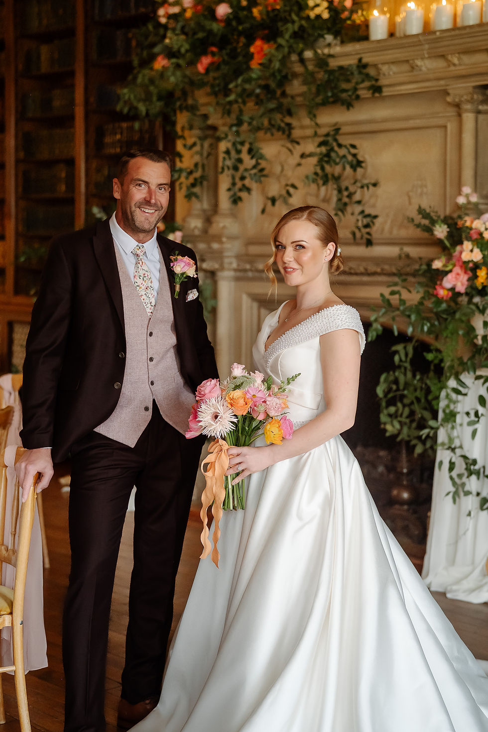 Smiling bride and groom in a candlelit hall, bride holding colorful bouquet, with flowers and greenery in the background.