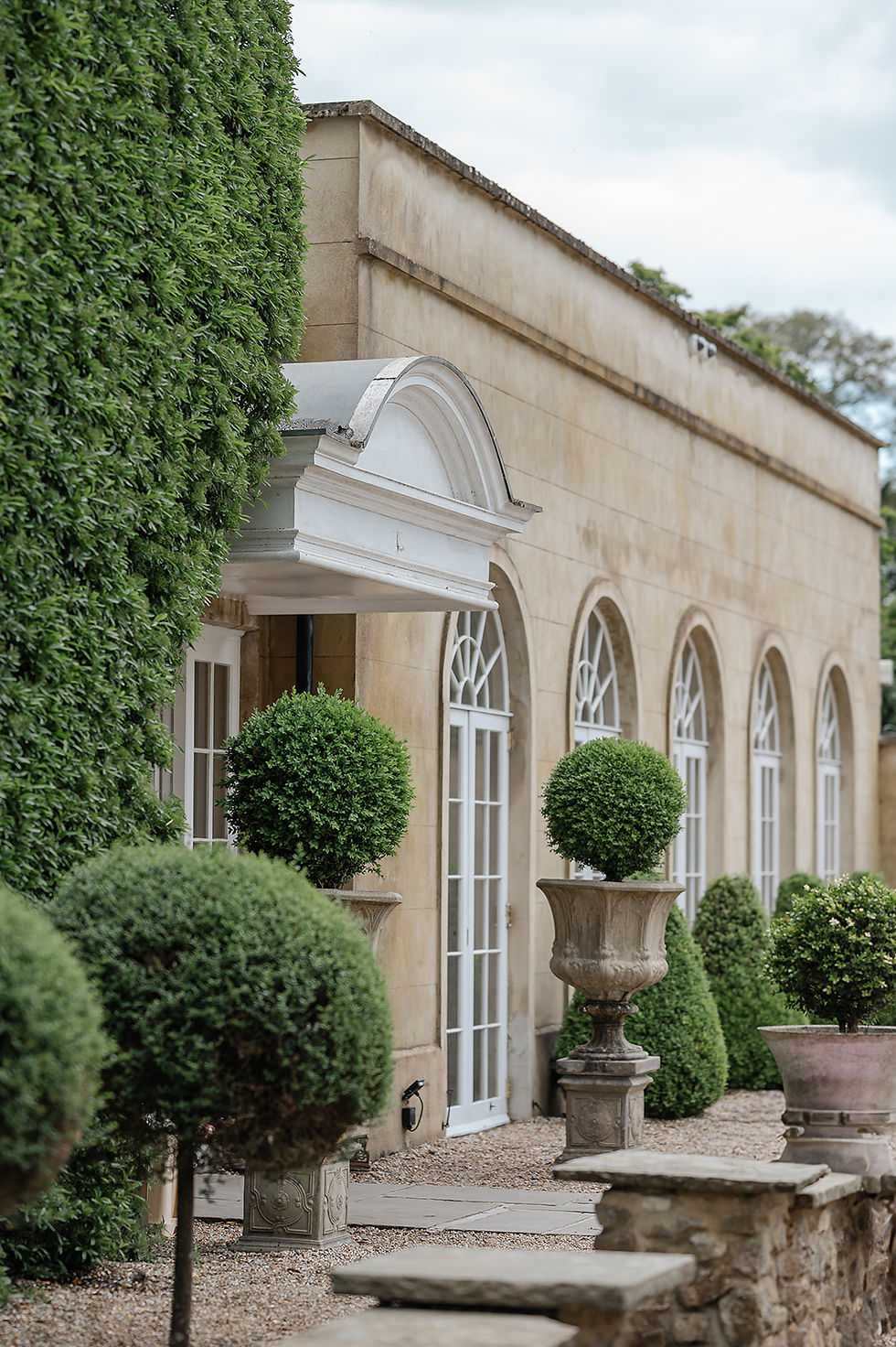 Elegant stone courtyard at Northbrook Park. arched windows and manicured topiary in urns, beside a tall green hedge.