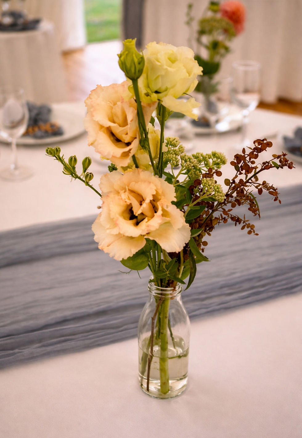 A glass vase holds peach roses with greenery, set on a white table with a light gray runner. Blurred table setting in the background.