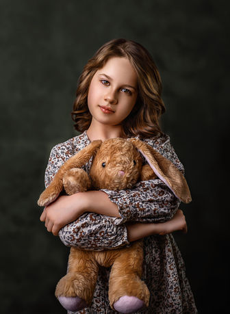 A young girl holds a stuffed bunny and stares at the camera against a dark green background in a fine art portrait capturing emotion and innocence.