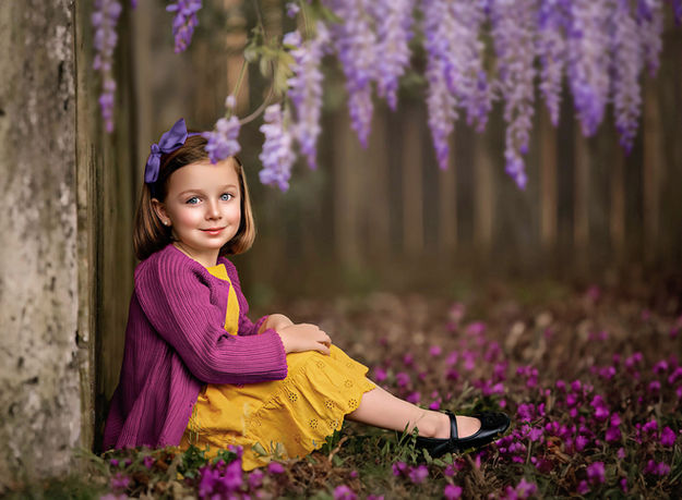 A little girl in a yellow dress and purple sweater sits against a wooden fence, with purple flowers hanging in the background in a fine art portrait filled with charm and softness- captured by Atlanta photographer