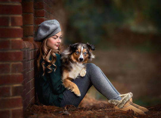 A young girl leans against a brick wall, holding an Australian Shepherd puppy on her lap, captured in profile in a fine art portrait.