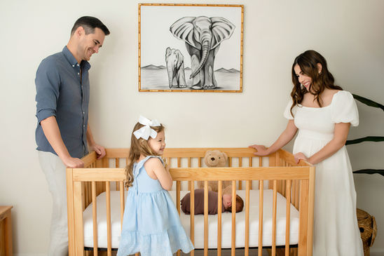Parents and toddler smiling around a crib while newborn baby sleeps inside.