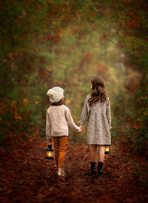 Two little girls walk hand in hand through a forest with soft fall colors, their backs to the camera, in a fine art portrait filled with warmth and nostalgia.