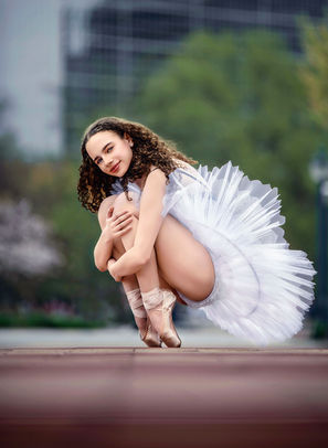 A dancer in a white pancake tutu kneels en pointe, holding her knees, set against an urban backdrop that contrasts elegance with grit.