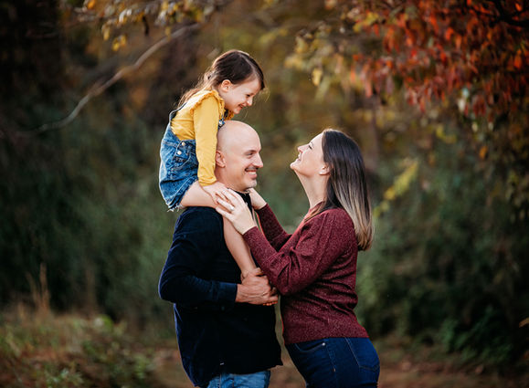 Little girl sitting on her dad's shoulders in an outdoor fall setting, with colorful autumn leaves in the background.