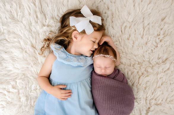 Toddler girl kissing her newborn sister’s forehead while the baby sleeps.