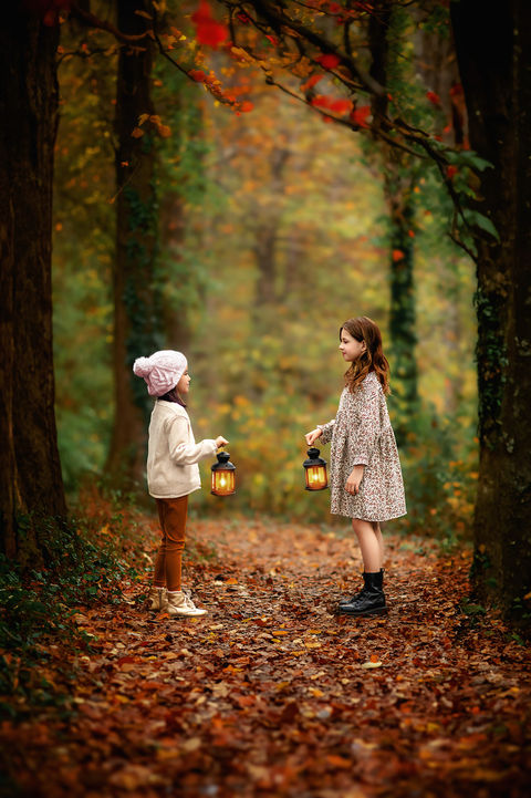 Two young girls stand in a forest with soft fall colors, facing each other while holding small lanterns in a fine art portrait that evokes warmth and wonder.