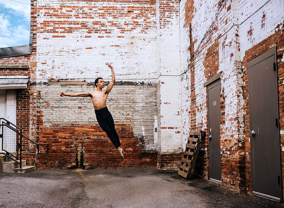 A male dancer performs a dynamic leap against a brick wall, showcasing strength, agility, and expressive movement.
