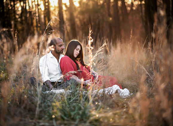 Pregnant woman sitting in a field of golden tall grass, with her partner sitting behind her, both enjoying a peaceful moment together.