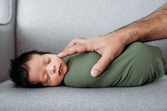 A newborn baby boy in a green wrap lays down peacefully as his dad’s finger softly touches his cheek