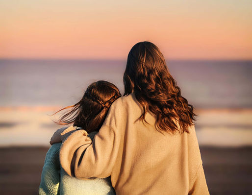 Back view of two sisters embracing, watching ocean sunset.