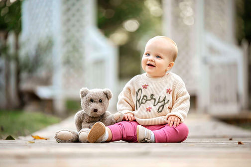 A one-year-old baby girl sits outdoors, smiling at something with a teddy bear next to her, capturing a sweet and joyful moment - captured by Atlanta baby photographer