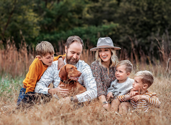 Family of five with three young boys and a brown dog sitting in a field of tall grass, looking at each other during a family session in Atlanta, Georgia