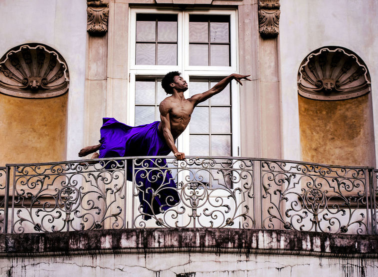 A male dancer in a flowing purple skirt executes a stunning jump on the balcony of a mansion, blending strength and elegance in midair.