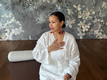 Frau in weißem Outfit, Hand auf Brust, sitzt auf Yoga-Matte. Hintergrund mit floralen Mustern. Ausdruck entspannt, bunte Ohrringe.