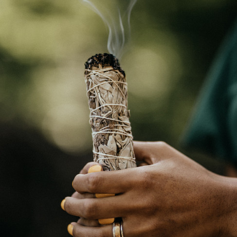 Hände halten ein brennendes Salbeibündel, aus dem Rauch aufsteigt. Hintergrund ist unscharf grün. Fingernägel sind gelb lackiert.