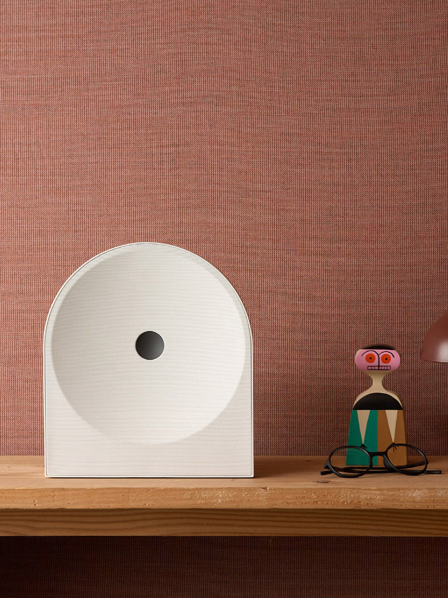 bafle de sonido samsung, estante de madera con objeto blanco redondeado, figura decorativa colorida y gafas sobre fondo rosa texturizado.