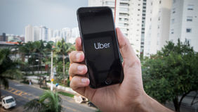 Mano sosteniendo un móvil con la app Uber abierta, frente a edificios altos y árboles en una calle urbana.