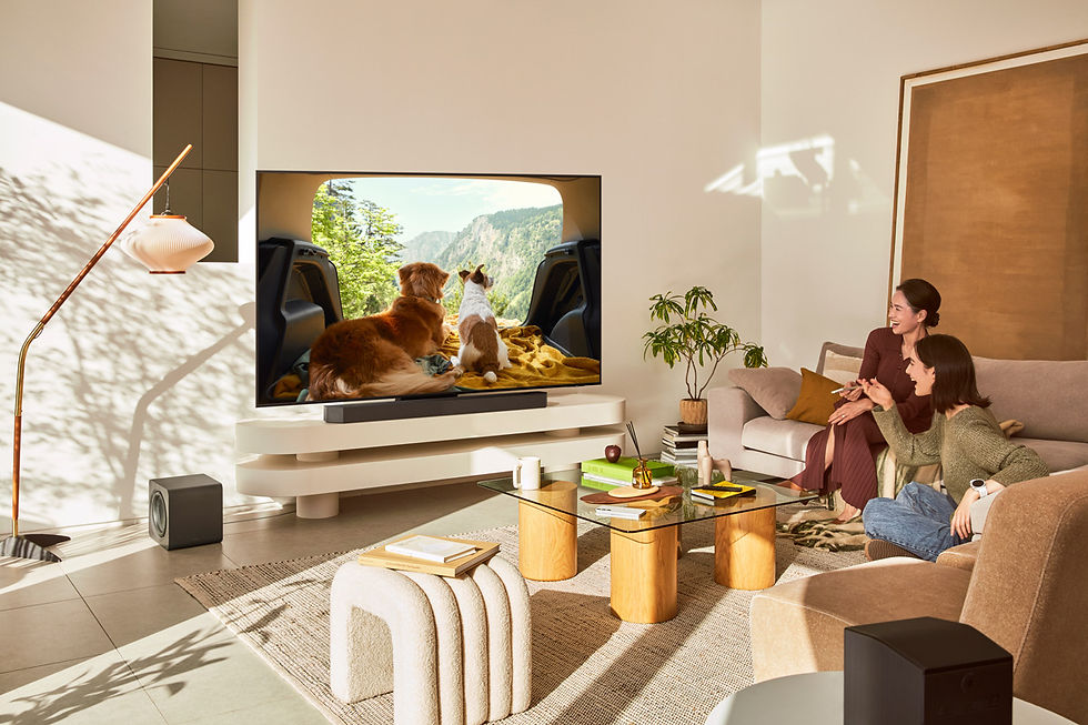 Dos mujeres en un sofá miran TV; en televisor samsung en pantalla, un perro y un gato frente a un paisaje montañoso en una sala luminosa y acogedora.