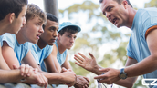 Coach passionately speaks to four young athletes in blue shirts outdoors, conveying focus and teamwork. Trees blur in the background.
