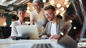 Three people collaborate at a table, smiling. One holds a paper, another types on a laptop. Modern office with warm lighting.