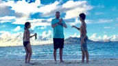 Craig Zuber with his children Aspen and Zach on a beach in Hawaii, Craig Zuber in blue shirt between two kids, facing the ocean and mountains under a bright cloudy sky.