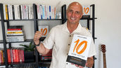 Business coach and author Craig Zuber holds an advance copy of The 10-Year Letter, smiling in front of bookshelves with a guitar and raised fist in the background.