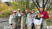 A group of people in fishing gear smile by a river with autumn trees. They wear colorful hats and jackets. A logo "CZ" is visible in the corner. Craig Zuber enjoying time outdoors with friends — blog about the power of disconnecting and reconnecting.