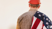 Worker in plaid shirt and red hard hat draped in an American flag, facing a bright, neutral background. Mood is patriotic.