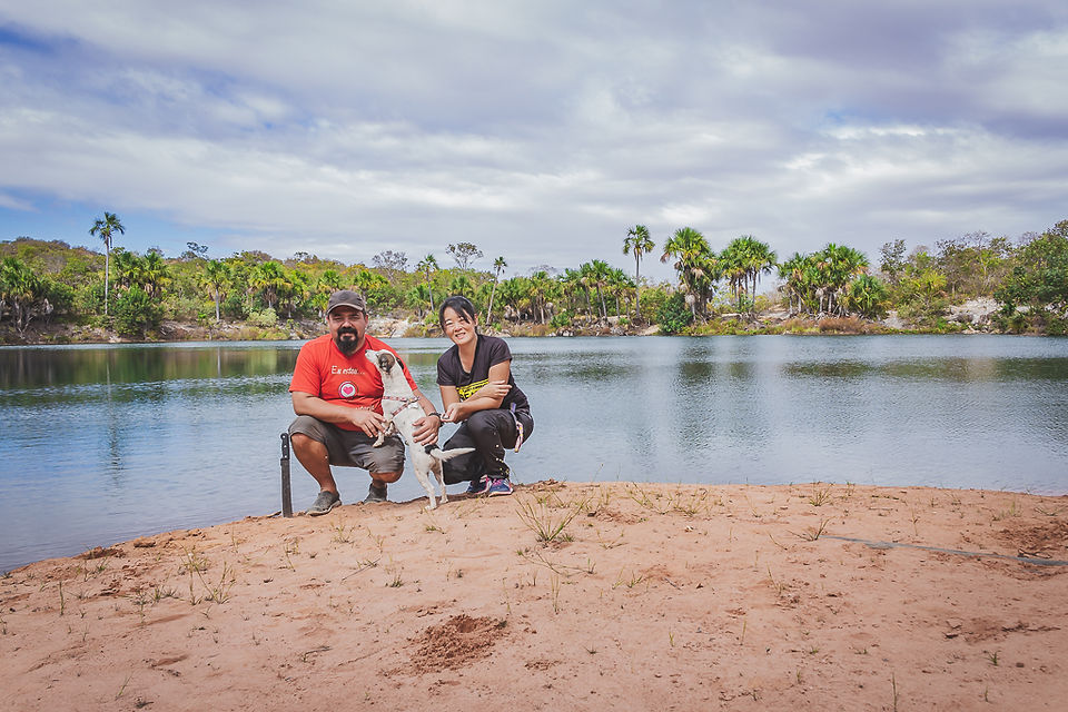 O casal do Viagem Kombinada, Cristiano Cazarotto, Sandra Doyama e a cachorrinha Mel na beira da Lagoa Feia em Coração de Jesus, MG. Eles estão agachados em uma faixa de areia. Atrás deles há a lagoa e ao fundo, a vegetação predominante de coqueiros contornando a lagoa.