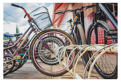 Em um ângulo visto mais de baixo avista-se à direita uma estrutura metálica em semi-arcos para o estacionamento de bicicletas. Na frente, a roda da primeira bicicleta emoldura as outras rodas das bicicletas mostrando vários círculos e aros alinhados.