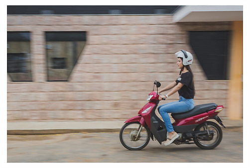 Uma moto pilotada por uma moça aparece com efeito de movimento, com o fundo mais borrado. Em uma moto vermelha à direita da imagem, uma moça com longos cabelos castanhos-escuro, veste sapatilha rosa claro, calça jeans, blusa preta e um capacete branco.