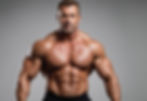 Muscular man posing shirtless against a gray background, showcasing defined muscles. His expression is confident and intense.