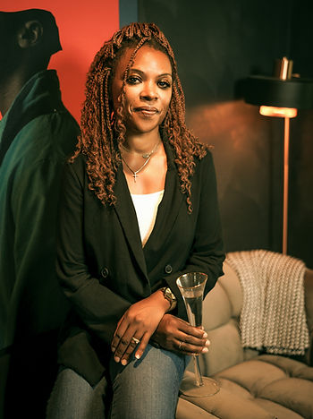 Amira Davis stands confidently next to a modern velvet armchair, holding a champagne flute. She wears a black blazer, white top, jeans, and pink heels. The room is dimly lit with a contemporary, moody aesthetic, and a large artistic portrait hangs behind her. She looks directly at the camera with a calm, grounded expression.