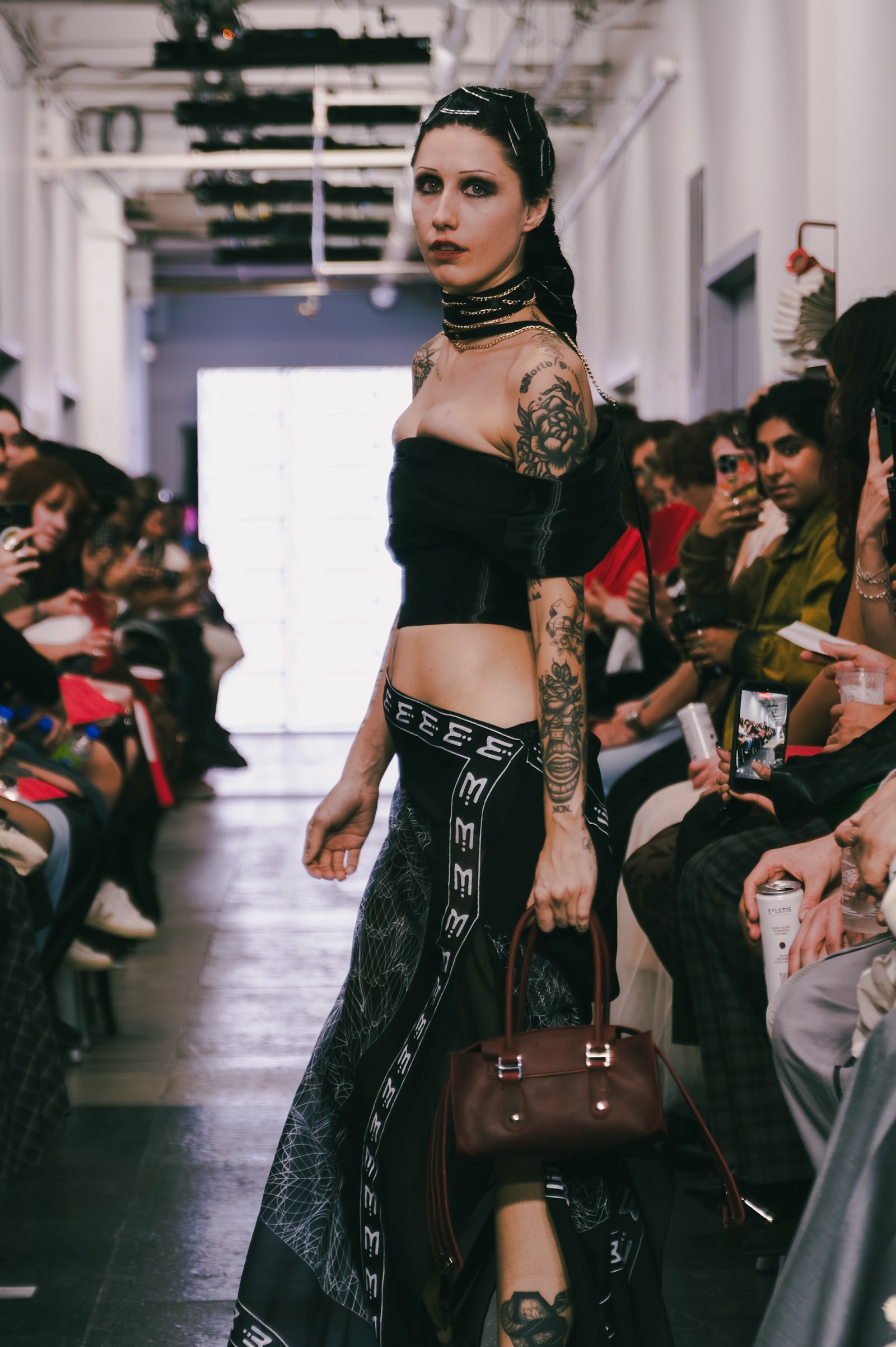 A tattooed model turns on the runway in a black cropped top and dark patterned skirt, carrying a brown bag.